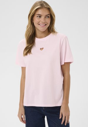 Femme aux longs cheveux blonds souriante, portant un t-shirt rose clair à manches courtes avec une petite découpe en forme de cœur près du col et un jean bleu foncé.