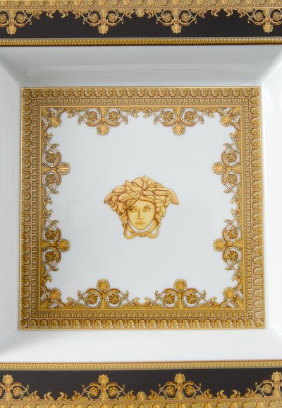 Kvadrātveida porcelāna šķīvis ar zelta un melnu izsmalcinātu apmali, centrā atrodas zelta Medūzas seja. Gluda virsma.
