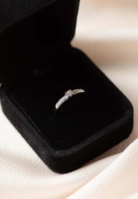 Zilveren ring met een centrale diamant en zijkanten, gepresenteerd in een zwart fluwelen ringdoos op een zachte stoffen achtergrond.