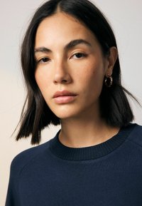 Mörkblå sweatshirt med ribbad krage, bärs av en person med kort mörkt hår och enkla hoopörhängen, mot en ljus bakgrund.