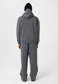 Grijze hoodie met een grote capuchon en drop shoulders, gecombineerd met bijpassende losse broeken met een achterzak. Geplaatst tegen een neutrale achtergrond.