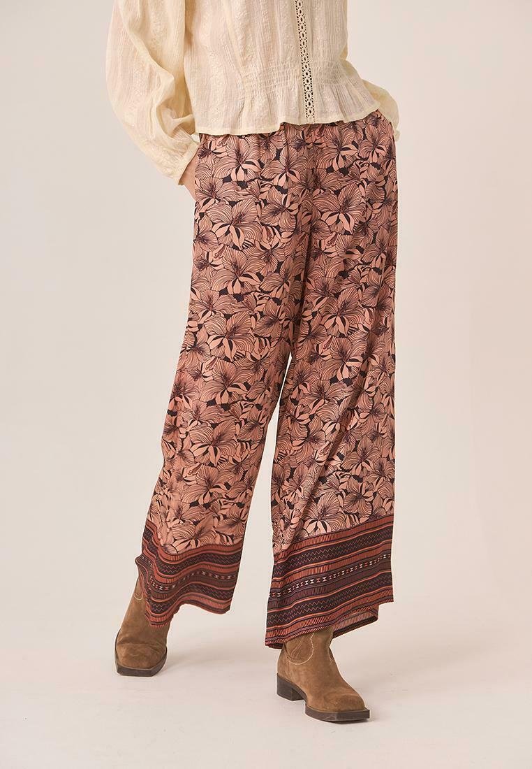 Persona con pantalones de pierna ancha de estampado floral y dobladillo a rayas, combinados con botas de ante marrón hasta el tobillo y una blusa texturizada color crema.