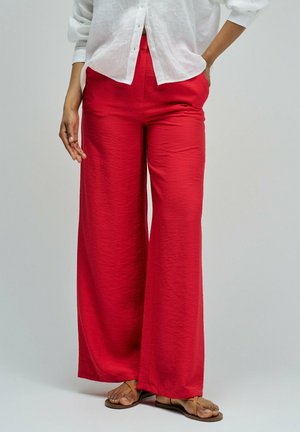 Camisa blanca de manga larga combinada con pantalones rojos de pierna ancha y talle alto, y sandalias planas color beige sobre un fondo neutro.