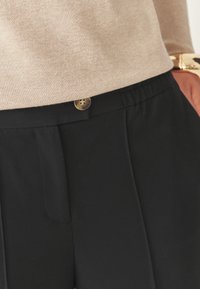 Pantaloni neri con una texture liscia, dotati di chiusura con bottone, vita elastica e pieghe frontali, parzialmente coperti da una blusa beige.