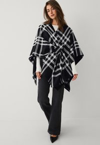 Sort og grå ternet poncho lavet af blødt stof, med et løst draperet design og trekantet snit. Parret med mørke jeans og hæle.