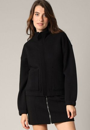 Veste noire zippée avec col montant, deux poches avant et un tissu texturé. Associée à une mini-jupe noire avec une fermeture éclair à l'avant.