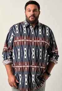 Hombre con barba vestido con una camisa de manga larga con un estampado, con el puño arremangado, mano izquierda en el bolsillo, de pie contra un fondo claro y liso.