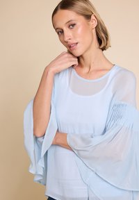 Lyseblå bluse med gjennomsiktig stoff, utsvingte ermer og rund hals. Har samlet detaljering på ermene og en glatt tekstur.