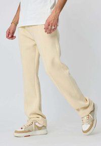 Pantalons de sudation beige, fuselés, en tissu doux avec des coutures visibles. Portés avec des sneakers clairs et multifacettes avec des accents orange.