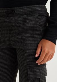 Harmaa houndstooth-kuvioinen collegehousut, joissa on joustava vyötärö ja nyöritys. Varustettu taskuilla ja sivussa cargo-tyylinen tasku.