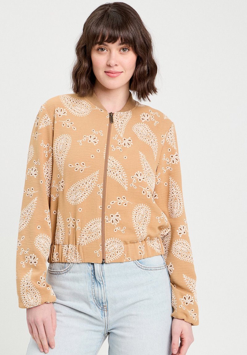 Kahki bomber jacket, jossa on valkoisia paisley-kuvioita, ribbed-kaulus ja vetoketjukiinnitys. Pehmeä kangas, lyhennetty leikkaus ja elastinen vyötärö.