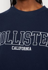 Granatowa bluza z prążkowanym, okrągłym dekoltem. Na froncie wyraźne białe napisy „HOLLISTER” nad „CALIFORNIA” w kontrastujących czcionkach. Miękki materiał.