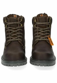 Botas de cuero marrón con punteras redondas, suelas de goma gris, cordones beige en contraste y una etiqueta naranja. Presentan ojetes reforzados y acentos cosidos.