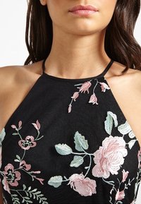 Haut halter noir avec broderies florales dans des nuances de rose et de vert, à la coupe ajustée et avec des bretelles délicates. Détail en tissu texturé visible.