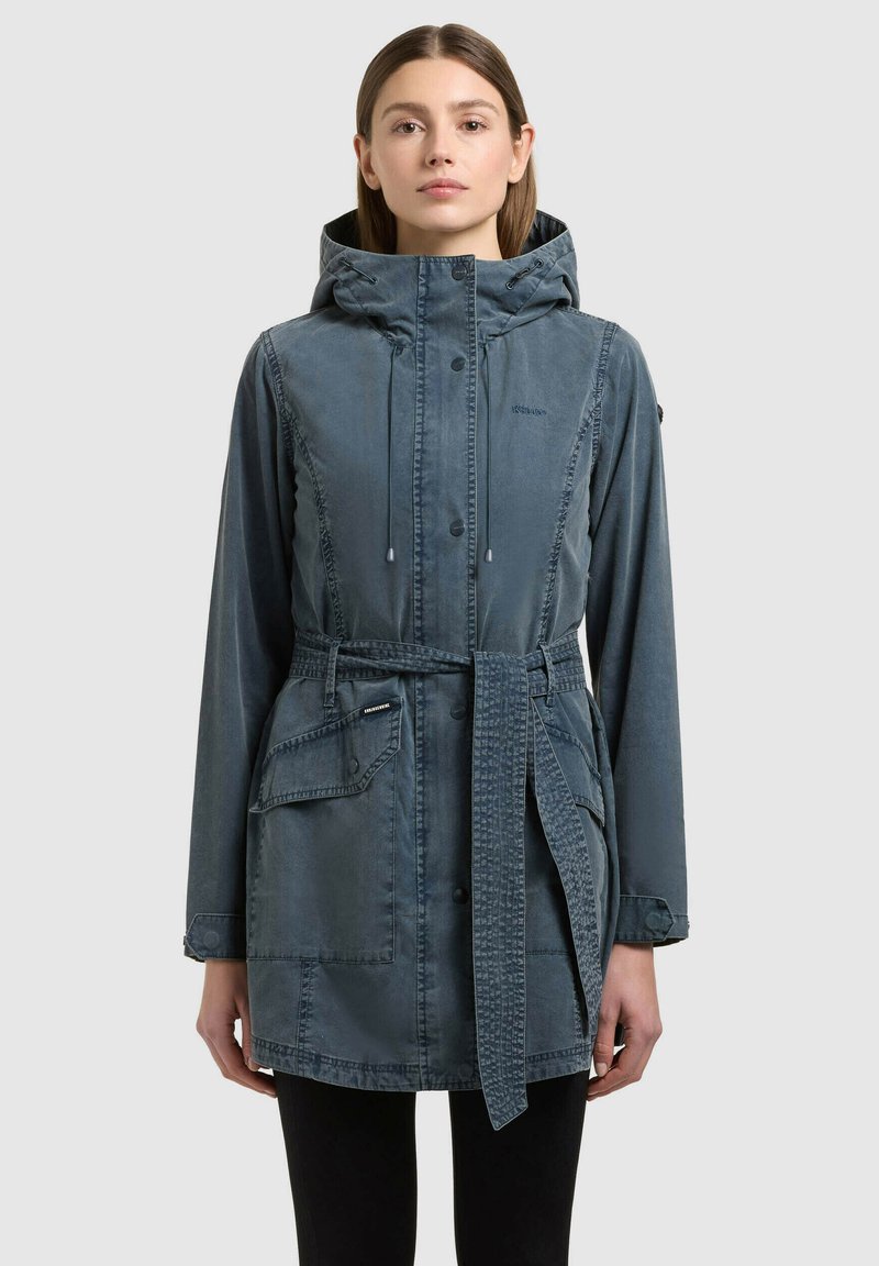 Femme portant une veste à capuche bleue mi-longue avec boutons-pression à l'avant, ceinture ajustable, poches à rabat et manches longues, debout devant un fond uni.