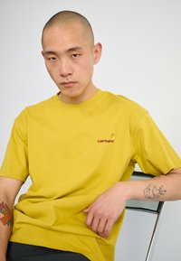 Geel katoenen t-shirt met korte mouwen en een ronde hals, voorzien van een klein rood geborduurd logo aan de linkerkant. Effen textuur.