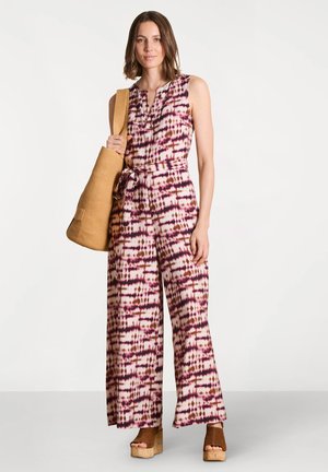 Femme portant une combinaison sans manches rose et marron à motif avec une ceinture assortie, tenant un sac fourre-tout tressé beige et portant des sandales à plateforme marron.