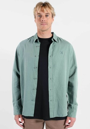 Chemise vert clair à boutons avec une texture douce, comportant un logo sur la poitrine et des manches longues, portée sur un t-shirt noir.