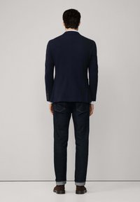 Marineblazer mit einem Schlitz, kombiniert mit dunklen Jeans, hochgekrempelten Ärmeln und braunen Lederschuhen, von hinten betrachtet.
