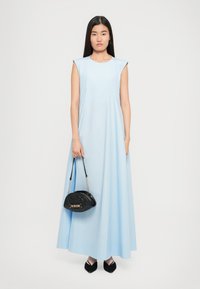 Γυναίκα σε αμάνικο, ανοιχτό μπλε, μακρύ φόρεμα, που κρατά μία μαύρη τσάντα με επένδυση και φορά μαύρες γόβες με αιχμηρή μύτη, στέκεται μπροστά σε άσπρο φόντο.