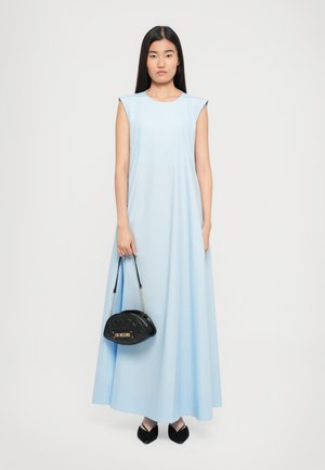 Woman in a sleeveless light blue floor-length dress holding a black quilted handbag and wearing black pointed-toe shoes, standing against white background.