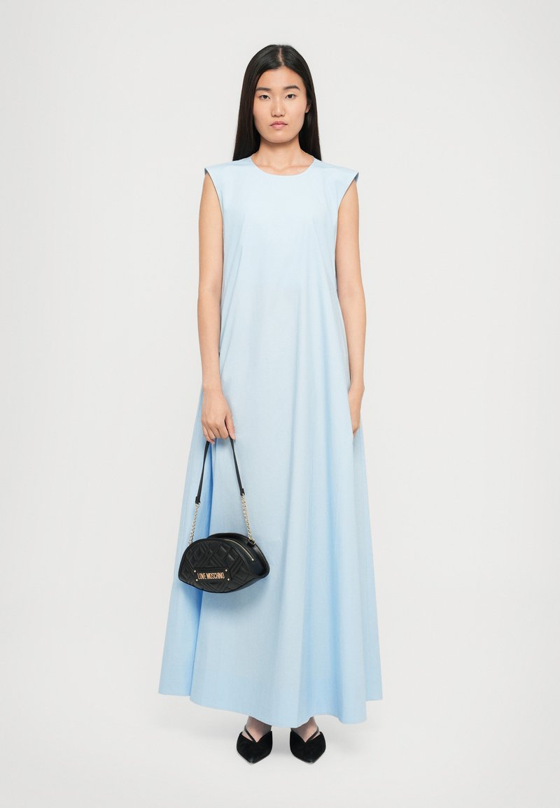 Sieviete bez piedakšiem, tērpta gaiši zilos garajos svārkos, tur melnu kvadrātveida somiņu un valkā melnas kurpes ar garo purngalu, stāv uz balta fona.