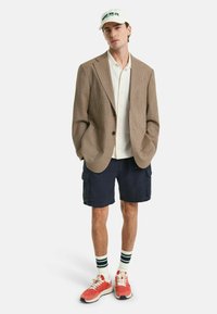 Braun kariertes Blazer über einem weißen Hemd, marineblaue Cargo-Shorts, rote Sneakers und weiß-grün gestreifte Socken, gekrönt mit einer weißen Kappe.
