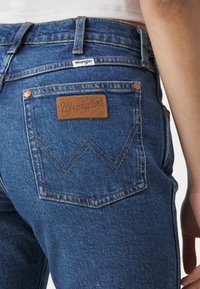 Jeans de mezclilla azul con cintura alta, que cuentan con una etiqueta de cuero marrón en el bolsillo trasero, costuras en contraste y un diseño clásico de cinco bolsillos.