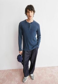 Langärmeliges, geripptes marineblaues Henley-Shirt mit Drei-Knopf-Leiste, kombiniert mit locker sitzender, dunkler Jeanshose. Graue Sneaker und eine marineblaue Mütze in der Hand.