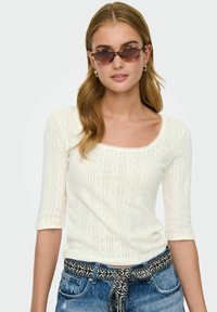 Crèmekleurig langemouwen top met een ronde halslijn, voorzien van verticale strepen en textuurpatronen, gecombineerd met blauwe denim shorts en een patroongordel.