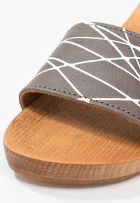 Sandal en bois à bout ouvert avec une sangle grise ornée de motifs géométriques blancs et de coutures visibles.