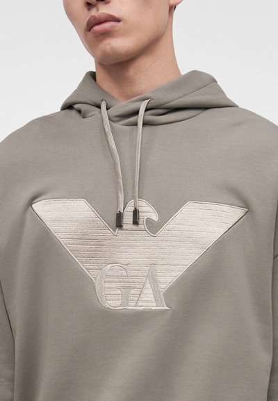 Gros plan d'une personne portant un sweat à capuche gris avec un grand logo aigle brodé et les lettres GA sur la poitrine.