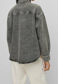 Veste en jean grise surdimensionnée avec un col pointu, des poches latérales et un ourlet effiloché. Présente des coutures visibles et une coupe décontractée.