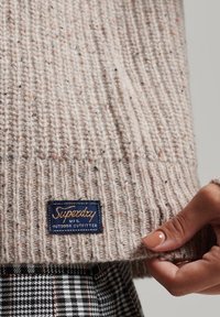 Pull tricoté beige clair avec une texture côtel ée et des flocons multicolores. Comprend une étiquette bleue avec "Superdry" en lettres dorées.