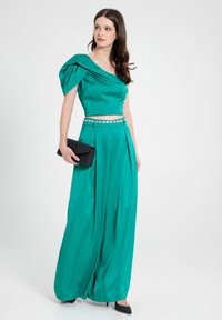 Türkises Satin-Kleid mit einem schulterfreien Design, plissiertem Rock und verziertem Gürtel; kombiniert mit einer schwarzen Clutch und schwarzen Absätzen.