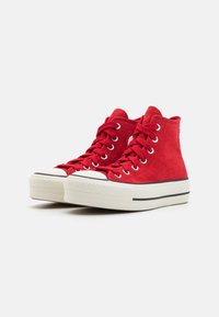 Czerwone zamszowe trampki typu high-top z białymi gumowymi podeszwami i czarnymi paskami, z okrągłymi metalowymi oczkami i czerwonymi sznurówkami.