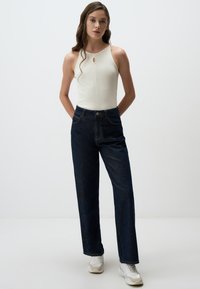 Canotta bianca a costine con dettaglio a chiave, abbinata a jeans skinny blu scuro a vita alta. La modella indossa sneakers bianche. Design semplice e aderente.