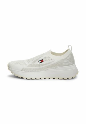 Zapatilla blanca tipo slip-on con parte superior de punto, suela gruesa, dibujo texturizado y un pequeño logo rectangular con la bandera en el lateral.