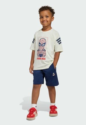 T-shirt beige chiaro con grafica di Spider-Man, abbinato a pantaloni corti blu navy. Sneakers rosse con tre strisce bianche. Capelli ricci e espressione sorridente.