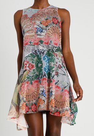 Vestito senza maniche, fluido con motivi floreali rosa, rossi, blu e verdi su uno sfondo chiaro, indossato da una persona in piedi davanti a uno sfondo semplice.