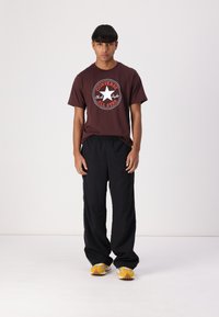 T-shirt marron à manches courtes avec un logo Converse blanc et rouge, associé à un pantalon noir ample et des chaussures de sport jaunes.