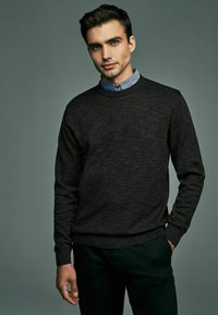 Un pull ras du cou gris foncé en tricot, doté de manches longues et de poignets côtelés. Porté au-dessus d'une chemise à col bleu clair.