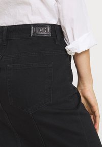 Svarta denimjeans med en synlig ficka och en etikett som läser "DIESEL." Texturen verkar slät, med en figurnära passform. En vit t-shirtärm är delvis synlig.