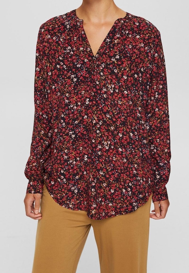 Femme portant un chemisier rouge à fleurs à manches longues avec un col en V, associé à un pantalon marron clair, sur un fond uni.