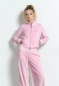 Młoda kobieta z blond włosami w wysokim kucyku, ubrana w jasnoróżową, welurową bluzę na zamek i pasujące szerokie spodnie, stojąca z rękami w kieszeniach.