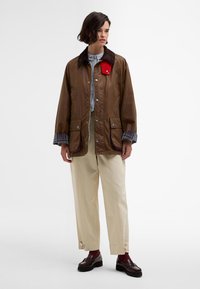 Brown outer jacket with a contrasting red collar, snap buttons, and pockets. Layered with a checked inner fabric. Paired with beige pants.