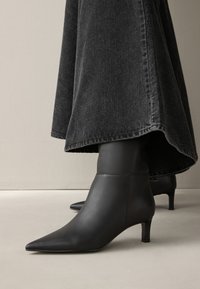 Bottes courtes noires à bout pointu avec talons kitten portées sous une longue jupe en denim gris foncé sur un sol clair.
