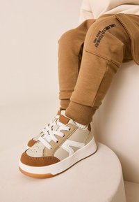 Beige och vita sneakers med bruna accenter, med vadderad tungspets, texturerad ovandel och vita snören. Kombinerat med bruna sweatpants.