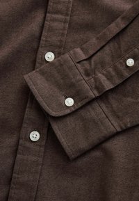 Brun, texturerad skjorttyg med knappärmar och vita knappar. Har en klassisk krage och snygga sömnadsdetaljer.