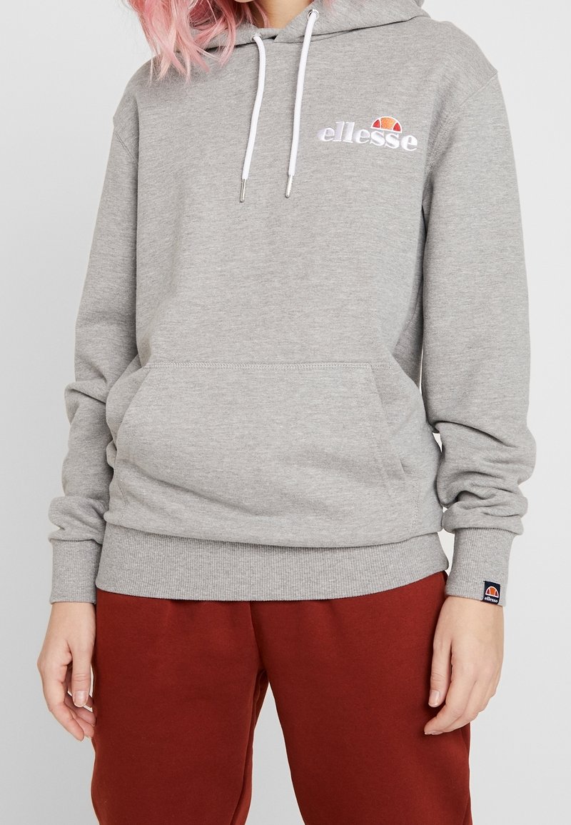 Personne portant un sweat à capuche gris ellesse avec poche avant et un pantalon rouge, debout devant un fond clair uni.
